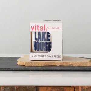 Puede incluir: Caja blanca con texto rojo y negro que dice "vital industries" y "LAKE HOUSE" en texto azul. La caja dice "HAND POURED SOY CANDLE" en la parte inferior.