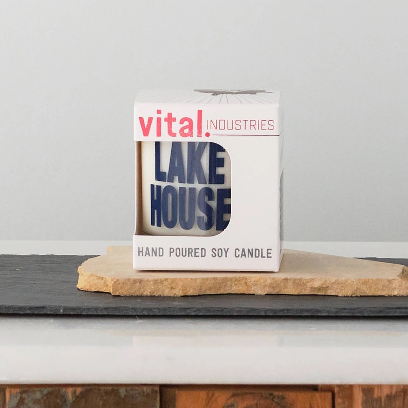 Puede incluir: Caja blanca con texto rojo y negro que dice "vital industries" y "LAKE HOUSE" en texto azul. La caja dice "HAND POURED SOY CANDLE" en la parte inferior.
