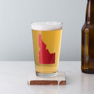 May include: A clear glass beer mug with a red outline of the state of Idaho on the side. The mug is filled with a light-colored beer and is sitting on a brown and white coaster.