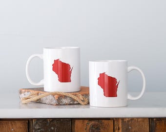 May include: Two white ceramic mugs with a red outline of the state of Wisconsin. The mugs are displayed on a wooden surface with a rope accent. The mugs have a simple, clean design.