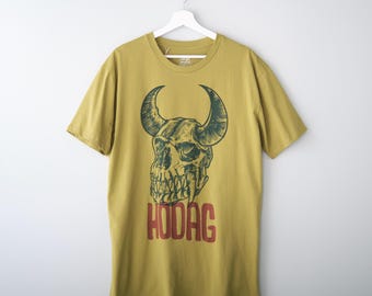 May include: A mustard yellow short-sleeve t-shirt featuring a dark green graphic illustration of a horned skull. Below the skull, the word HODAG is printed in bold red capital letters. The shirt is displayed on a white plastic hanger.