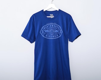 May include: A royal blue short-sleeve t-shirt featuring a light blue graphic on the chest. The graphic is an oval emblem containing the text "EXPLORE THE NORTHWOODS WISCONSIN" with a sun, water, and pine tree illustration inside the border.