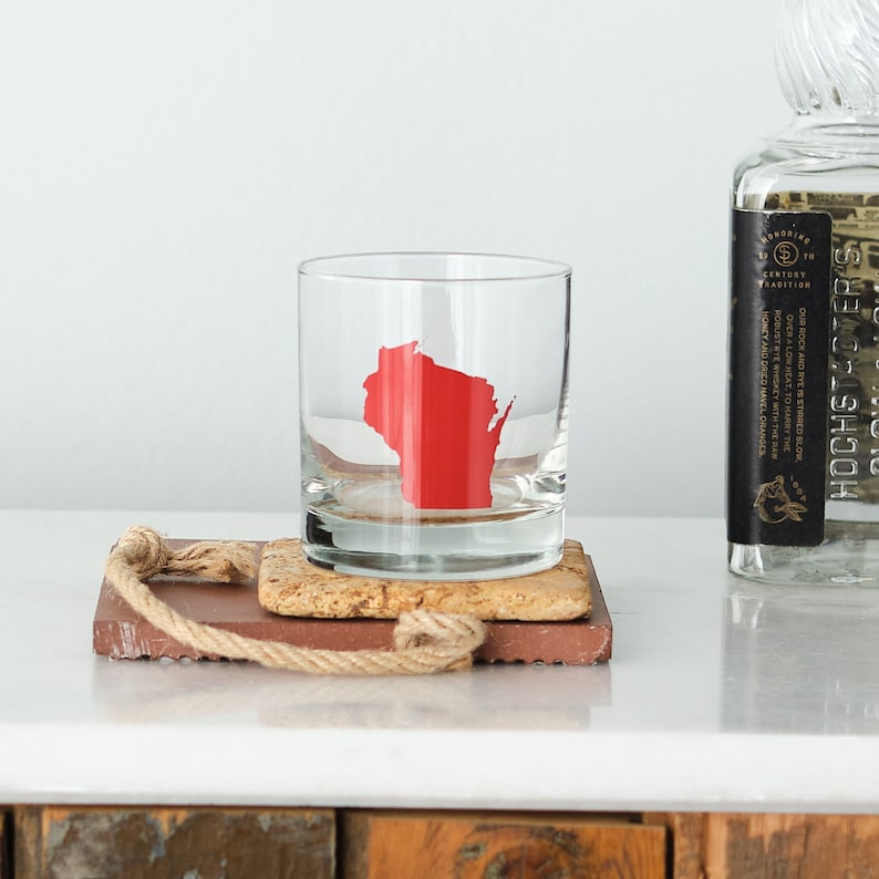 May include: A clear glass rocks glass with a red outline of the state of Wisconsin. The glass is sitting on a brown coaster with a rope tied around it.