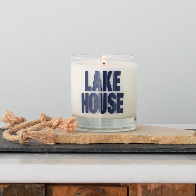 Puede incluir: Una vela blanca en un tarro de vidrio transparente con una etiqueta azul "LAKE HOUSE". La vela est&aacute; encendida y se encuentra sobre un posavasos de piedra marr&oacute;n.