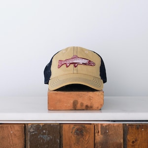 May include: Tan baseball cap with a navy blue mesh back and a maroon fish graphic. The cap is sitting on a brick. The hat has a curved brim and a structured front panel.