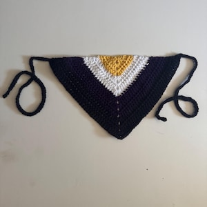 May include: A handmade crochet bandana in a triangular shape. The bandana features a color block design with a black base, a white stripe, and a gold-colored center. Black ties are attached to the corners for securing the bandana.