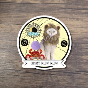 May include: A sticker featuring a cat wearing a lion's mane, holding yarn, and a crab. The design includes zodiac symbols and the text "CRABBY MEOW MEOW" on a banner. The background is yellow with sun-like rays.