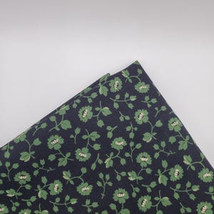 May include: Folded fabric with a dark navy blue background and a repeating floral pattern. The flowers are light green with off-white centers, and the leaves are a darker green. The fabric is likely cotton or a similar material.