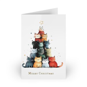 May include: A white greeting card featuring a watercolor illustration of a Christmas tree made of cats. The cats are various colors, and a gold star sits atop the tree. The words "Merry Christmas" are printed in gold.