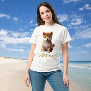 以下が含まれることがあります： 柴犬の子犬のグラフィックがプリントされた白いTシャツ。子犬は茶色と白で、ふわふわの尻尾と警戒した表情をしています。「JUST BE」の文字が子犬の下に金色でプリントされています。カジュアルな普段着です。