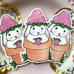 May include: Three white bunny stickers with pink strawberry hats and green leaves. Each bunny is sitting in a brown pot with a green leaf sticking out of the side. The bunnies are holding a small pink strawberry in their paws.