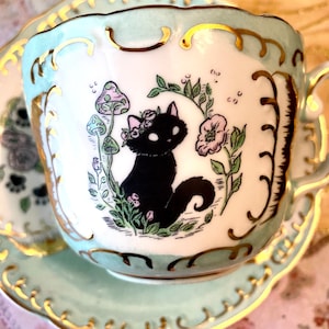 May include: A decorative teacup and saucer set. The cup and saucer are light green with gold trim. The cup features a black cat illustration surrounded by flowers and mushrooms. The teacup has a handle.