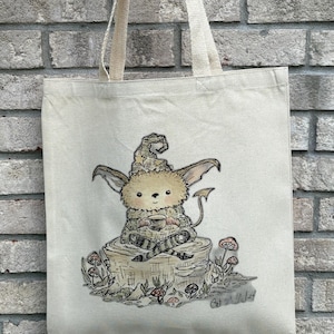May include: Cream-colored tote bag featuring a whimsical illustration of a furry creature with a hat, holding a cup, and sitting on a tree stump. The design includes mushrooms and foliage, with the artist's signature in the bottom right corner.