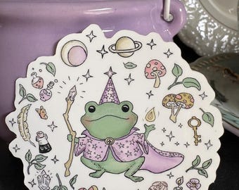Puede incluir: Una pegatina caprichosa con una rana verde mago con un bastón y una capa rosa, rodeada de setas, estrellas y botellas de pociones. La pegatina tiene un fondo blanco con un borde festoneado.