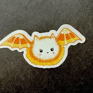 May include: A sticker featuring a cute, cartoon-style bat with a fluffy white body and orange and yellow wings. The bat has small black eyes and rosy cheeks. The sticker is die-cut with a white border.