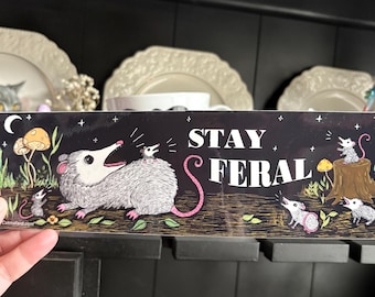 May include: A black sticker featuring the text "STAY FERAL" and illustrations of opossums, mushrooms, and foliage. The opossums are depicted in various poses, with a large opossum and several smaller ones. The sticker has a whimsical, cartoonish style.