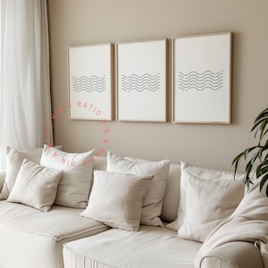 Op de afbeelding: Drie ingelijste prints met een minimalistisch ontwerp van zwarte golvende lijnen op een witte achtergrond. De prints hangen aan een beige muur boven een witte bank met witte kussens.