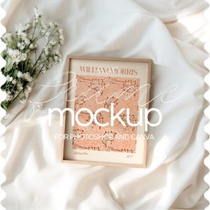 May include: A mockup of a framed print with a floral design. The frame is light brown wood and the print is a vintage style with a red and orange floral pattern. The text "William Morris" is visible at the top of the print. The frame is sitting on a white fabric surface.
