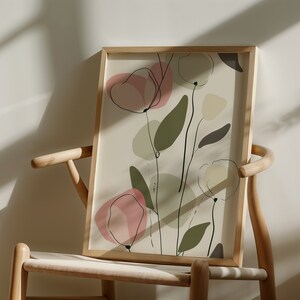 May include: A framed abstract art print with a beige background and pink, green, and brown floral shapes. The print is in a wooden frame and is leaning against a light wood chair.