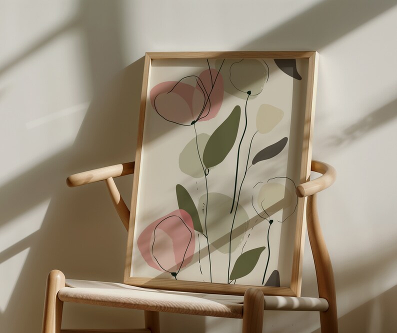 May include: A framed abstract art print with a beige background and pink, green, and brown floral shapes. The print is in a wooden frame and is leaning against a light wood chair.