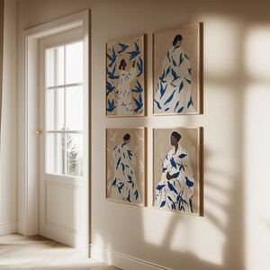 May include: Four framed art prints featuring abstract figures in white robes adorned with blue leaf-like patterns. The artwork is set against a beige background, with wooden frames. The prints are displayed on a white wall near a door.
