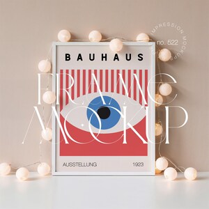May include: White framed Bauhaus art print with a red, white, and blue eye design. The print features the word "BAUHAUS" at the top, "AUSSTELLUNG" at the bottom, and the year "1923". A string of white globe lights is draped around the frame.