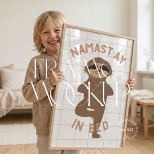Könnte beinhalten: Ein gerahmter Druck mit einem Cartoon-Faultier in einer Yoga-Pose, mit den Worten "NAMASTAY IN BED" in einer weichen, erdigen Farbpalette. Der Druck wird von einer Person gehalten, die die Größe und das Design des gerahmten Kunstwerks zeigt. Der Rahmen ist aus hellem Holz.