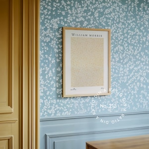 May include: A framed print with the text "William Morris" in a light wood frame, hanging on a blue wall with a white floral pattern. The print features a yellow and white floral design. A circular sticker with the text "Ratio Dina Ratio No. 213 Dina Ratio" is on the wall below the print.