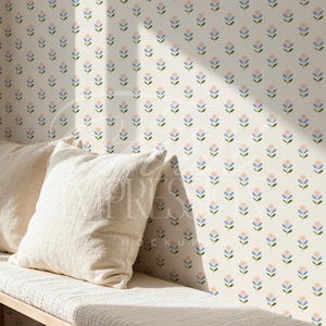 May include: A cream-colored wall with a repeating floral pattern in blue, pink, and green. Two cream-colored pillows rest on a white bench. The scene is lit by sunlight.