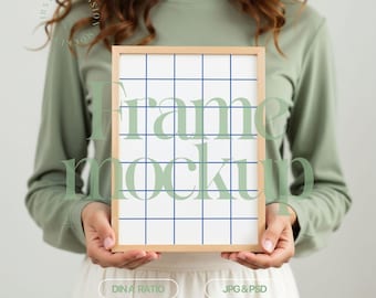 Op de afbeelding: Een persoon houdt een houten frame mockup vast met een witte achtergrond en een blauw rasterpatroon. Het frame wordt voor een witte muur gehouden. De tekst "Frame Mockup" is in lichtgroene kleur over het rasterpatroon geschreven.