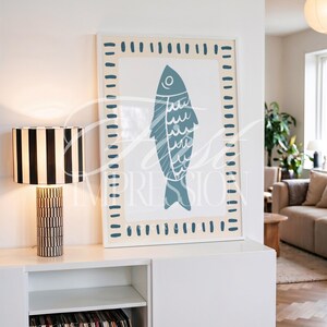 May include: A framed art print featuring a blue fish illustration on a cream background, surrounded by a dotted border. A black and white striped lamp sits on a white cabinet. The lamp has a cylindrical base and a rectangular shade.