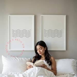 May include: A woman is sitting in bed reading a book. She is wearing a white vest top and has long dark hair. The bed is white and has a white pillow. There are two framed prints on the wall behind her, each with a black and white wave design. The text on the prints reads "A RATIO DIN A RATIO DIN".