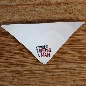 May include: White triangular fabric with the text "cupid's wing man" printed in black with a red and white plaid pattern.