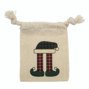 May include: A small, beige drawstring bag with a festive elf leg design. The elf legs are red and black plaid, with green elf shoes and a green and black plaid hat. The bag has a natural rope drawstring.
