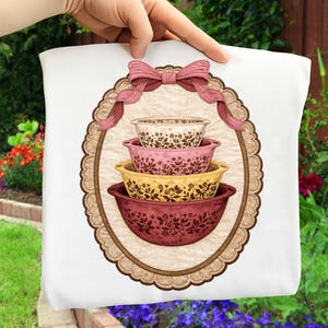 Peut inclure: Un coussin blanc avec un motif décoratif de quatre bols à mélanger superposés, dans les tons crème, rose, jaune et bordeaux. Les bols présentent un motif floral et sont encadrés dans un ovale marron avec un nœud rose.