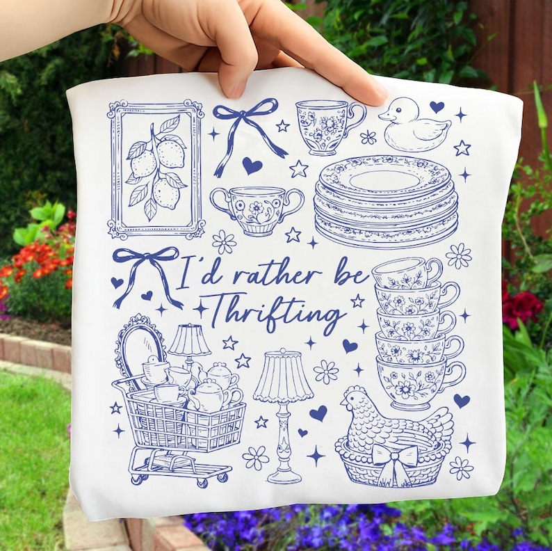 May include: White tote bag with a blue illustration of various vintage items, including teacups, plates, a shopping cart, and a chicken in a basket. The text "I'd rather be thrifting" is also included.
