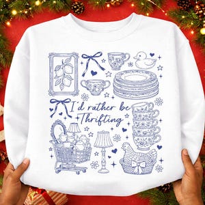 May include: White sweatshirt with a blue graphic design featuring various thrifted items. The design includes teacups, plates, a duck, a shopping cart, lamps, and the text "I'd rather be thrifting."