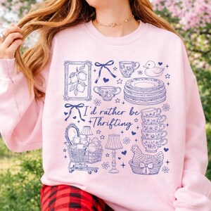 May include: A light pink sweatshirt with a blue graphic design featuring various vintage items like teacups, plates, and a shopping cart. The text "I'd rather be thrifting" is also included in the design.