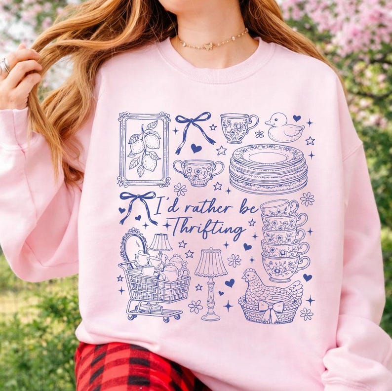 May include: A light pink sweatshirt with a blue graphic design featuring various vintage items like teacups, plates, and a shopping cart. The text "I'd rather be thrifting" is also included in the design.
