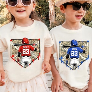 May include: Two children wearing cream-colored t-shirts with baseball-themed graphics. One shirt features a red baseball player with the name "Emily" and the number "23." The other shirt displays a blue baseball player with the name "John" and the number "23."