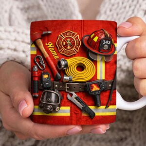 May include: A white ceramic mug featuring a firefighter's tools and gear design. The design includes a red axe, helmet, and a yellow hose, along with the text "Fire Rescue". The mug is held by a person.