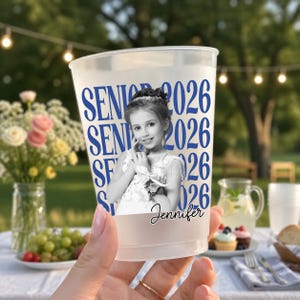 May include: A clear plastic cup with the words "SENIOR 2026" in blue. The cup features a black and white photo of a young girl and the name "Jennifer" in cursive. The cup is held in front of a blurred outdoor background.