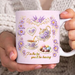 May include: A light pink ceramic mug with a floral design and the text "I'm the tea you'll be having." The mug features a teapot, teacup, and macarons. The design includes purple flowers and gold accents.