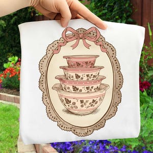 May include: White tote bag featuring a vintage illustration of four pink and cream-colored mixing bowls stacked inside an oval frame. The frame has a scalloped edge and a pink bow at the top. The bowls have a floral design.