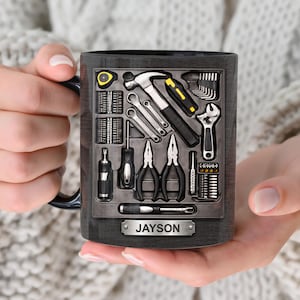 Puede incluir: Taza de café negra con diseño de caja de herramientas. La taza presenta una ilustración detallada de varias herramientas, incluyendo un martillo, llaves y alicates. El nombre "Jayson" se muestra en una placa metálica en la parte inferior del diseño.