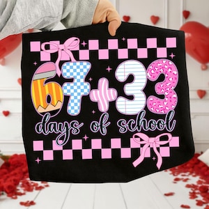 May include: Black tote bag with a colorful graphic celebrating 67 + 33 days of school. The design includes a pencil, checkered patterns, and the text "days of school" in a cursive font. Pink bows and stars complete the design.