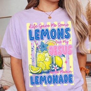 Può includere: T-shirt lilla chiaro con un design grafico. Il design presenta il testo "Life Gave Me Some Lemons But My Jesus He Be Makin Lemonade". Include un barattolo di limonata, limoni e una cannuccia.