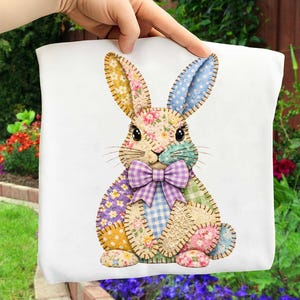 May include: A white square pillow with a patchwork bunny design. The bunny has floral, polka dot, and plaid patterns in yellow, blue, purple, and pink. It features a purple bow tie and black eyes. The pillow is held up by a hand.