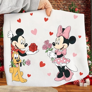 May include: A white blanket featuring Mickey Mouse, Minnie Mouse, and Pluto. Mickey Mouse is holding a gift, Minnie Mouse holds roses, and Pluto holds a rose. Pink and red hearts are scattered across the blanket.