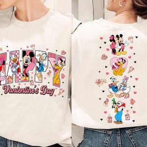 May include: Cream-colored sweatshirt featuring Disney characters and the text "Happy Valentine's Day." The front displays Minnie Mouse, Donald Duck, Mickey Mouse, and others. The back shows Mickey and Minnie Mouse, Pluto, Donald and Daisy Duck, and Goofy.